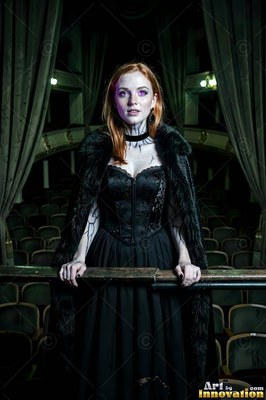 Wearing a black corset and fur cape, a pale-skinned woman stands at an old theater balcony rail