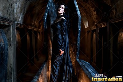 Standing in an open coffin, a pale woman in a black dress looks up in a stone crypt
