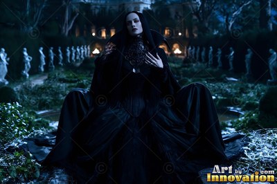 A woman with pale skin sits in a huge black gown in a dark garden lined with white statues