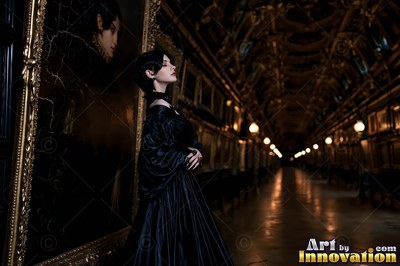 In a black silk gown, a pale-skinned woman is standing beside a gold framed painting in a long dark hall