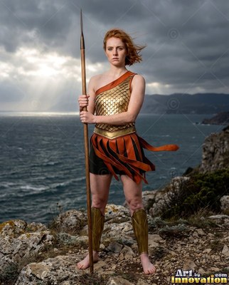 On a rocky cliff above the sea, a barefoot warrior woman in gold armor holds a spear