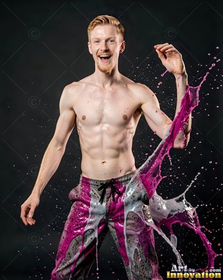 Laughing in studio light, a shirtless man is hit by splashes of pink and gray paint
