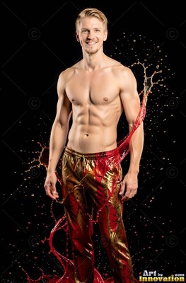 Standing in a dark studio, a smiling shirtless man is splashed with red liquid