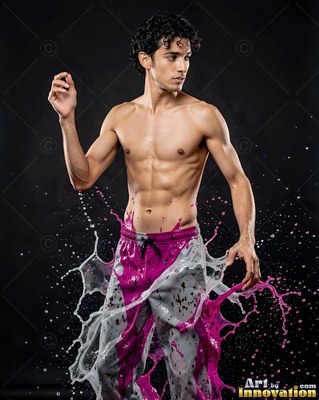 A shirtless man is standing as pink and white liquid splashes up around his pants against a dark studio background