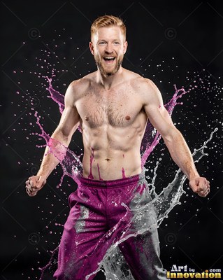 In a dark studio, a shirtless smiling man is hit with splashing pink liquid