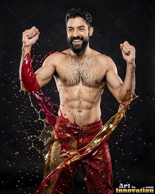 A shirtless man raises his fists as red and gold liquid splashes under bright studio light