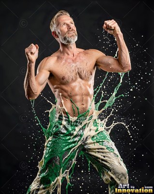 A shirtless man with a gray beard raises his fists in a dark studio as green and white paint splashes up his legs