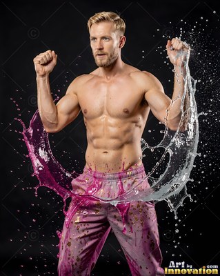 In a dark studio, a shirtless muscular man raises his fists as pink liquid and water splash