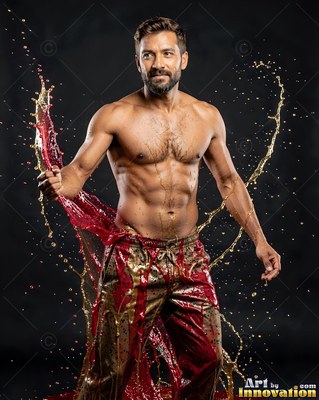 Standing in a dark studio, a shirtless man is splashed with red and gold water