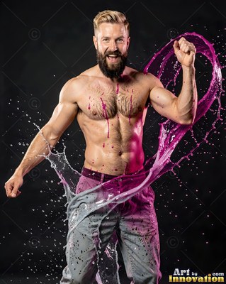 Wearing gray jeans in a dark studio, a shirtless man pumps his fist as pink water splashes
