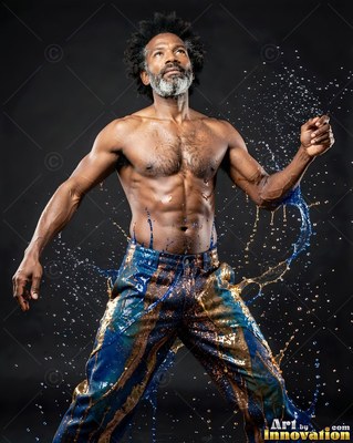 A gray-bearded shirtless man poses in a dark studio as blue and gold water splashes from his pants