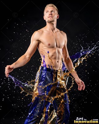 Standing in a dark studio, a shirtless man is splashed with blue and gold liquid