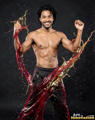 A smiling shirtless man raises his fists as red and gold water splashes around him