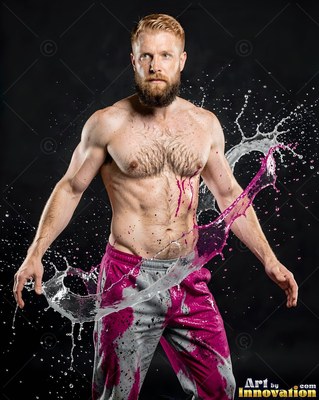 In a dark studio, a shirtless bearded man is standing as pink fluid splashes past him