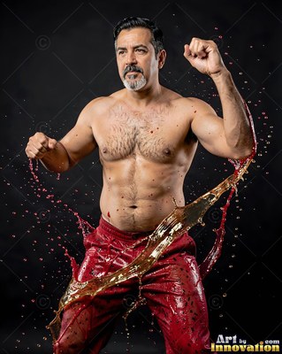 Fists raised in a dark studio, a shirtless man is splashed with red liquid