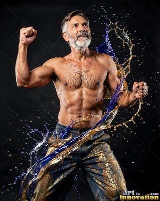 Raising his fists, a shirtless bearded man is splashed with blue and gold liquid