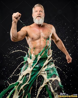 A shirtless bearded man pumps his fist as green and white paint splashes over him