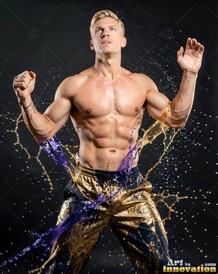 In a dark studio, a muscular blond man flexes as water splashes around him