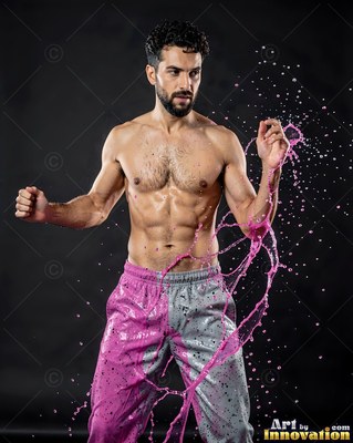 Posing in gray pants, a shirtless man is hit by a splash of pink liquid in a dark studio