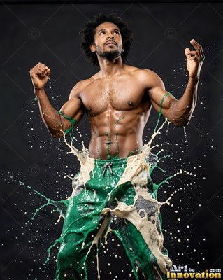 Wearing green sweatpants in a dark studio, a shirtless man raises his arms as white and green paint splashes