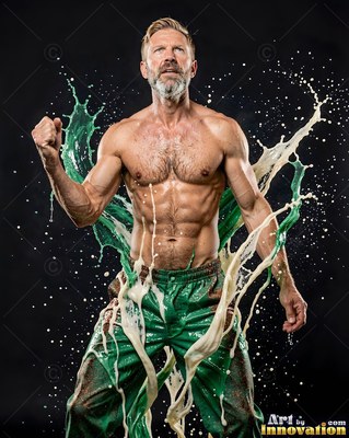 A shirtless muscular man in green pants pumps his fist as white paint splashes past him