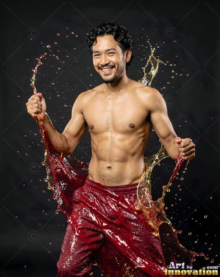 In a dark studio, a shirtless smiling man is splashed with red and gold liquid