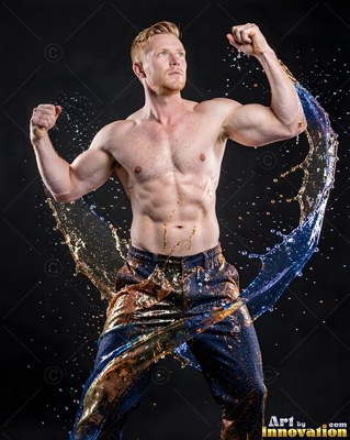 A shirtless man flexes his arms in a dark studio as splashes of blue and gold water curve around his body