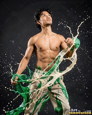 In a dark studio, a shirtless man looks up as green and cream paint splashes past