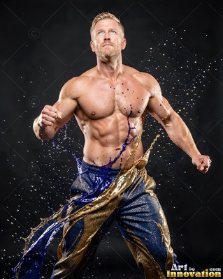 Standing in a dark studio, a muscular man is splashed with blue and gold liquid