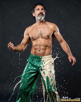 Standing shirtless, a muscular bearded man gets splashed with green and white paint