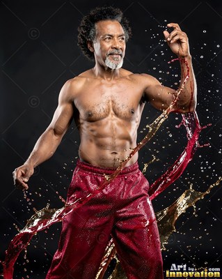A shirtless man poses with raised hands as splashing liquid catches bright light in red and amber hues