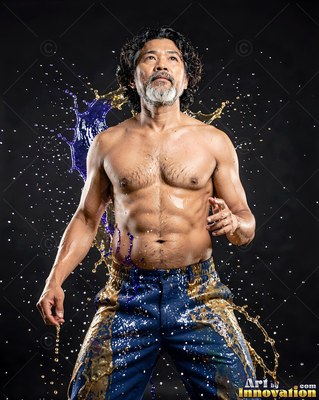 A gray-bearded shirtless man stands in a dark studio as blue and gold liquids splash behind him