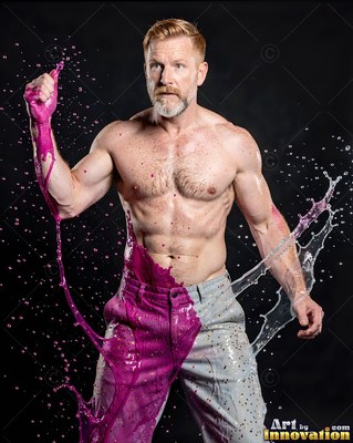 A shirtless bearded man poses as bright pink liquid splashes over him