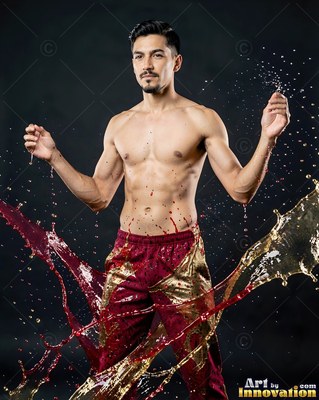 In patterned red pants in a dark studio, a shirtless man stands as red and gold liquid splashes past his legs