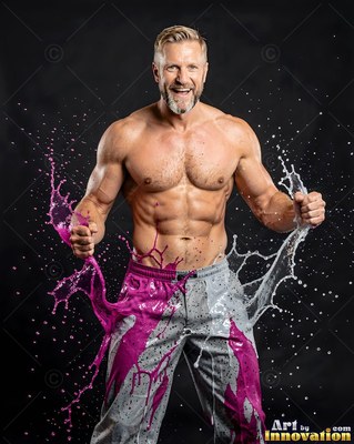 A shirtless muscular man laughs as bright magenta and white liquid splashes up his pants