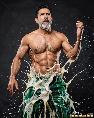 In a dark studio, a shirtless muscular man is splashed with creamy liquid around his waist