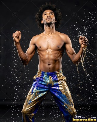 A shirtless man in shiny blue and gold pants stands as water splashes around him