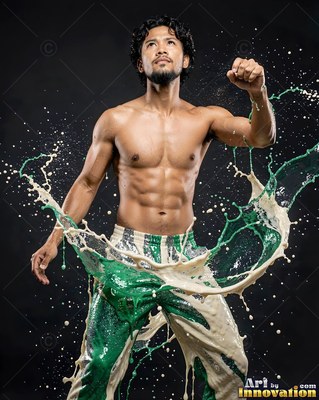 Standing with raised fist in a dark studio, a shirtless man is splashed with green paint