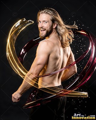 Long blond hair sweeps behind a shirtless smiling man ringed by red and gold water in a dark studio