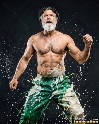 A muscular bearded man in green pants flexes as white liquid splashes around his waist