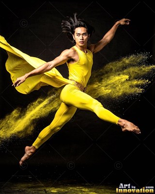 A man leaps across a dark studio in a yellow outfit trailing yellow powder