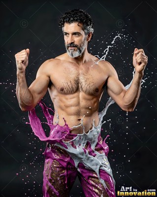 Gray-bearded shirtless man flexes his arms as pink and white paint splashes his waist in a dark studio