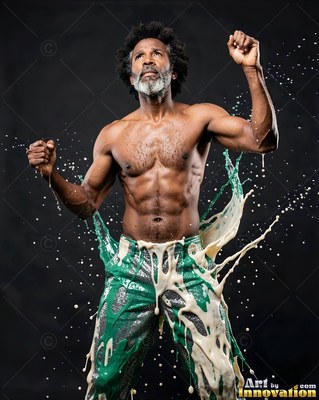 A shirtless man raises his fists as green and cream paint splashes over his pants