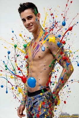 In a white studio, a smiling shirtless man is splattered with colorful paint droplets