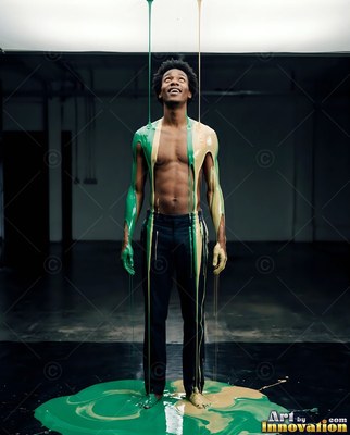 A shirtless man smiles up as green and tan paint pours over him in a dark room