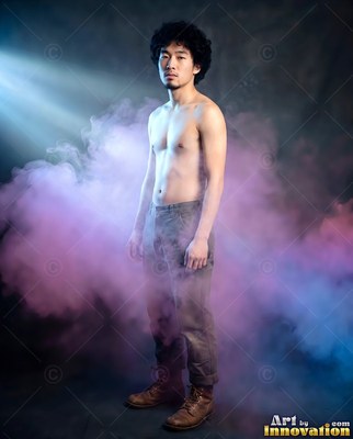 In purple smoke, a bare-chested man standing in gray trousers under a cool light beam