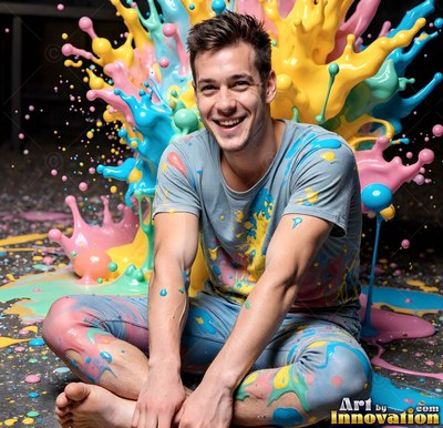 Wearing a paint-stained t-shirt on a dark floor, a smiling barefoot man sits as poured yellow and blue paint splashes behind him