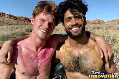 Smiling outdoors, two shirtless men with pink and yellow paint covering their chests pose together