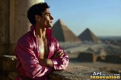 A man with an open pink shirt leans on a stone balcony facing the pyramids