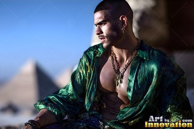 Wearing an open green soft silk shirt, a young man sits before the pyramids in bright sun